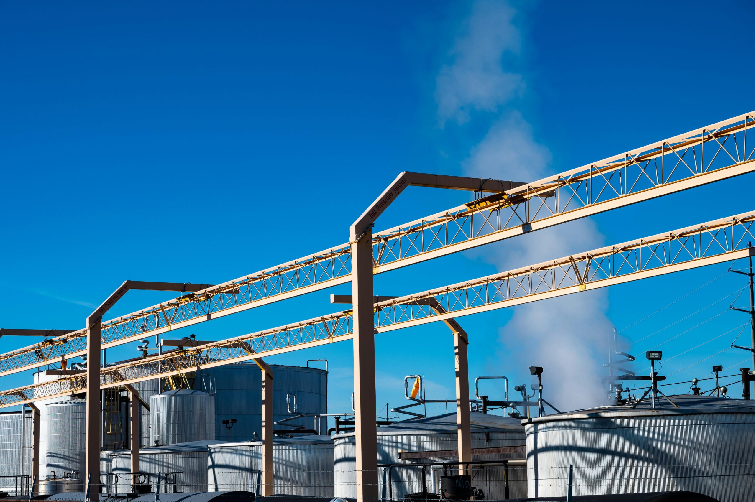 Wide view of an industrial plant with smoke rising from storage tanks against a clear blue sky.